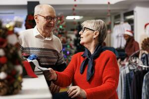 Cheerful elderly couple shopping at mall, looking to buy formal attire for christmas eve dinner outfit. Senior people checking ties from accessories box at clothing store, xmas decorations. photo