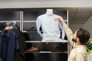 joven hombre leyendo rojo rebaja precio etiqueta mientras compras para ropa en negro viernes en Moda centro comercial, masculino cliente en pie en ropa Tienda despeje sección mirando a descontado camisa foto