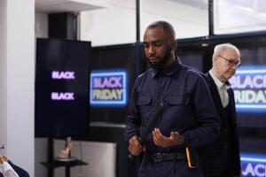 Serious African American man security officer in uniform patrolling shopping mall during Black Friday sales, ensuring crowd control, standing at clothing store entrance preventing shoplifting photo