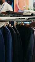 vertical vídeo hombre mirando a elegante chaquetas americanas en ropa almacenar, molesto a determinar Si materiales son cualitativo. cliente analizando vestidos en Moda boutique bastidores, decidiendo a comprar después hallazgo video