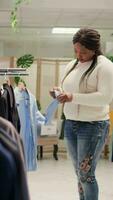 vertical vídeo mulher olhando às roupa formal camisas dentro roupas loja, tentando para determinar elas em forma dela. comprador analisando vestuário em moda boutique prateleiras, olhando para certo Tamanho roupas video