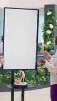 Vertical video Employee in shopping center fashion boutique using high tech screen to show woman clothes options to try on. Shopkeeper using led panel to help client visualize outfit combinations