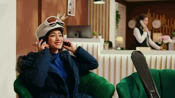 Asian person preparing to go sliding and sitting in lounge area at mountain ski resort, checking skis gear and headgear. Winter sport fan getting equipment ready for skiing down slopes. video