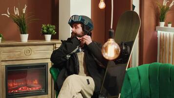 Athlete checking snowboard gear in lobby, sitting in lounge area and getting ready for snowboarding session on snowy mountain slopes. Young man with winter attire and helmet. Handheld shot. video