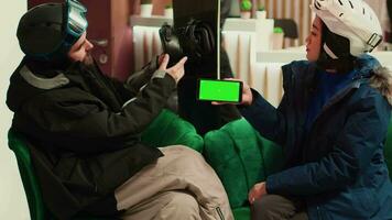 Woman holds smartphone with greenscreen while she talks to friend about winter sports at mountain ski resort. People equipped with skiing gear and protection, isolated mockup template. video