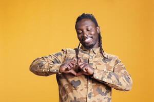 Smiling adult doing heart shape with hands on camera, promoting love in front of camera in studio over yellow background. African american excited lovely person expressing love feelings and gratitude photo