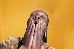 retrato de agotado africano americano persona cubierta boca con mano siendo cansado mientras posando en estudio en pie terminado amarillo antecedentes. hombre bostezando teniendo sobrecargado de trabajo expresión foto