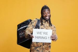 Takeaway delivery worker with thermal backpack holding job hire sign for courier position. African american man wanting perfect candidate for career opportunity in studio over yellow background photo