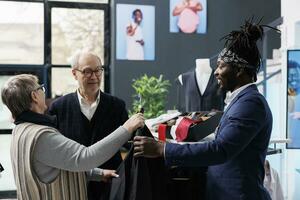 Old couple checking shirt material with boutique employee, buying trendy clothes for casual wear in clothing centre. Elderly customer looking at fashion collection in shopping centre. Fashion concept photo