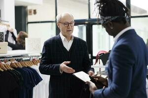 Elderly man asking worker for help with formal wear, african american employee checking merchandise inventory on tablet computer in showrrom. Senior customer looking at new fashion collection photo