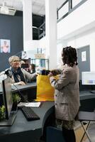 African american employee preparing bag with purchase for elderly client in modern boutique. Fashionable woman buying stylish casual wear in store, making electronic payment at pos terminal. photo