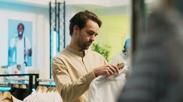 caucásico hombre mirando a de moda vestir en perchas, comprando moderno ropa en rebaja en compras centro. masculino cliente examinando Moda boutique mercancías, comercial actividad. Mano disparo. foto