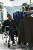 Arab man customer in wheelchair engaging with seller, discussing jackets available in clothing store. Client with disability examining apparel style and size while shopping for formal outfit photo