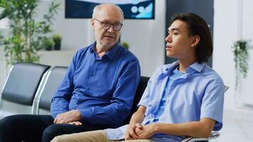 Patients sitting in waiting area lobby at hospital reception, preparing to start checkup visit with physician. Senior man discussing with disease symptoms with asian patient. Medicine service photo