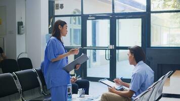 Assistant standing at hospital counter desk, writing medical expertise on clipboard. Asian patient calls the nurse to help him with insurance information during checkup visit in waiting room photo