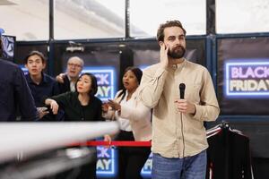 Young man TV news reporter standing with microphone against angry crazy crowd of shoppers waiting for discounts. Journalist correspondent making special reportage about Black Friday event photo