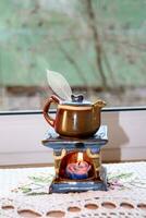Aroma lamp in the form of a teapot with a burning candle inside and an openwork leaf by the window, vertical photo