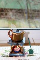 Aroma lamp in the form of a teapot with a burning candle inside and an openwork leaf and a burning incense stick by the window, vertical photo