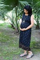 A pregnant Indian lady poses for outdoor pregnancy shoot and hands on belly, Indian pregnant woman puts her hand on her stomach with a maternity dress at society park, Pregnant outside maternity shoot photo