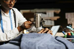 Close up of african american couturier using industrial sewing machine on fabric material in tailoring studio. Suitmaker cutting clothing garment piece, creating bespoke attire photo