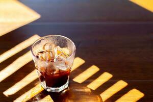 Cola in a glass with ice cubes on a wooden table with shadow photo