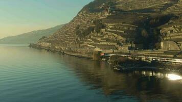 passager train va le long de Lac Genève rive à le coucher du soleil. réflexion dans l'eau. en terrasse vignobles de lavaux. Suisse. aérien voir. drone suit train video