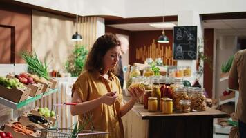 sonriente vegano mujer en especialidad cero residuos supermercado agregando abultar artículos con no contaminante embalaje a compras cesta, retrato. cliente en local barrio tienda comprando toxinas gratis nutritivo comida video