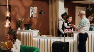 Hotel concierge looks at passport to complete check in forms at reception, senior man waiting at front desk. Tourist arriving in lobby with luggage, ready to register and start retirement holiday. video