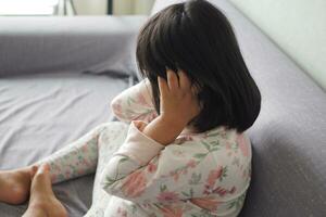 un trastornado niño niña cubrir su cara con mano sentado en sofá foto