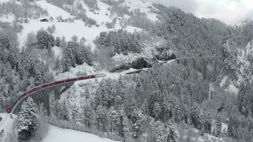 Aerial video of famous train of red color Glacier Express at winter, The train moves to the Landwasser Viaduct and tunnel, landmark of Switzerland, snowing, drone flies forward