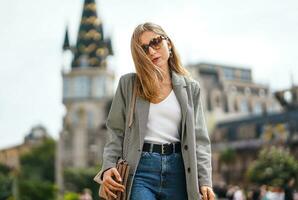 Tired girl walks through city. Beautiful architectural buildings. Woman wearing jacket and jeans with sun glasses walking around square. Sunny summer day. Blurred background. photo