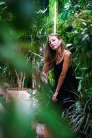 hermosa mujer en pie y mirando directamente a cámara. tropical plantas. joven mujer vistiendo negro vestir dentro invernadero. elegante y elegante moda. retrato de atractivo joven morena mujer. foto