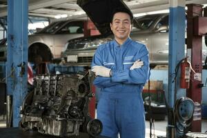 Portrait of Asian male mechanic supervisor in uniform, arms crossed and looking at camera near car engine at service garage, happy maintenance work, check and repair occupation in automotive industry. photo