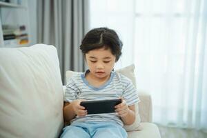 Asian child girl looking using and touch mobile phone screen on couch sofa. Baby smiling funny time to use mobile phone. Too much screen time. Cute girl watching videos while tv, Internet addiction. photo