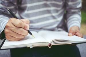 Close up of man's hands writing in spiral notepad placed on wooden desktop with various items photo