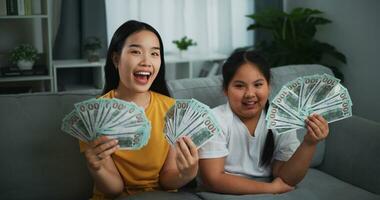 retrato de joven asiático mujer demostración dólar a cámara y sonriente en sofá en el vivo habitación a hogar. foto