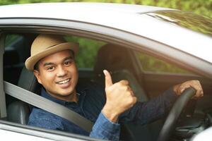 Portrait of happy smiling young asian man with fasten seat belts for safety travel on the road showing thumbs up while driving in his car. photo