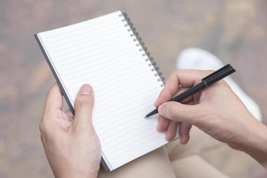 close up hand young man are using pen writing Record Lecture note pad into the book sitting on the chair outdoor. Blank book page copy  write a message. photo