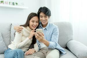Asian couple smiling and happily use smartphone to online shopping. Husband ready to pay birthday present for beautiful wife. Find accommodation online for honeymoon, App online, book flight tickets. photo