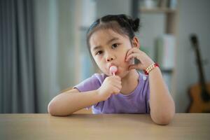 adorable Asia linda bebé niña jugando y comiendo con vistoso niño jugar arcilla piruletas en el de madera mesa en el vivo habitación a hogar. foto