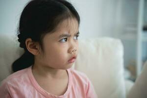 Asian kid girl upset lonely bullied little looking away feels abandoned abused, sad alone sitting on the sofa couch in living room at home, charity adoption concept. photo