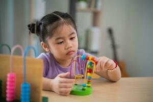 joven linda asiático bebé niña es aprendizaje el ábaco con de colores rosario a aprender cómo a contar en el mesa en el vivo habitación a hogar. niño bebé niña desarrollo estudiando concepto. foto