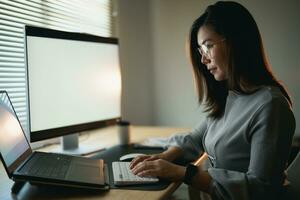 asiático mujer de negocios vistiendo traje y lentes mecanografía en teclado y trabajando ordenador portátil en de madera mesa a hogar. empresario mujer trabajando para su negocio a vivo habitación hogar. negocio trabajo hogar concepto. foto