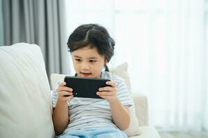 Asian child girl looking using and touch mobile phone screen on couch sofa. Baby smiling funny time to use mobile phone. Too much screen time. Cute girl watching videos while tv, Internet addiction. photo