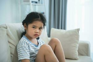 Asian kid girl upset lonely bullied little looking at camera feels abandoned abused, sad alone sitting on the sofa couch in living room at home, charity adoption concept. photo