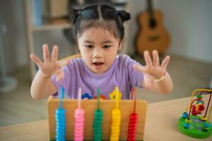 joven linda asiático bebé niña es aprendizaje el ábaco con de colores rosario a aprender cómo a contar en el mesa en el vivo habitación a hogar. niño bebé niña desarrollo estudiando concepto. foto