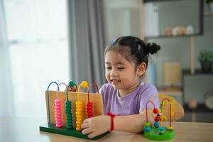 joven linda asiático bebé niña es aprendizaje el ábaco con de colores rosario a aprender cómo a contar en el mesa en el vivo habitación a hogar. niño bebé niña desarrollo estudiando concepto. foto