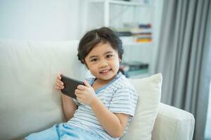 Asian child girl looking using and touch mobile phone screen on couch sofa. Baby smiling funny time to use mobile phone. Too much screen time. Cute girl watching videos while tv, Internet addiction. photo