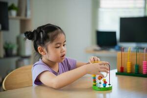 joven linda asiático bebé niña es aprendizaje el ábaco con de colores rosario a aprender cómo a contar en el mesa en el vivo habitación a hogar. niño bebé niña desarrollo estudiando concepto. foto