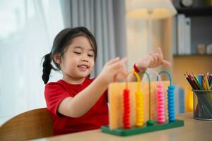 Young cute Asian baby girl wearing red t-shirt is learning the abacus with colored beads to learn how to count on the table in the living room at home. Child baby girl development studying concept. photo